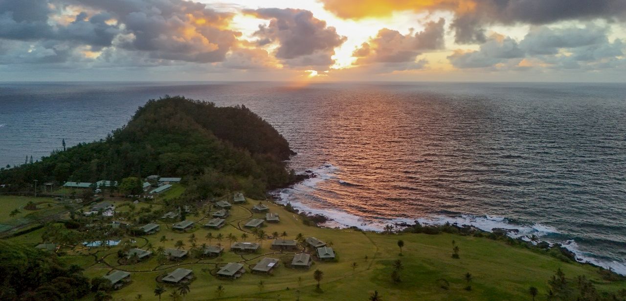 A beachfront resort by a green headland watches the sun set over the ocean with glowing clouds.