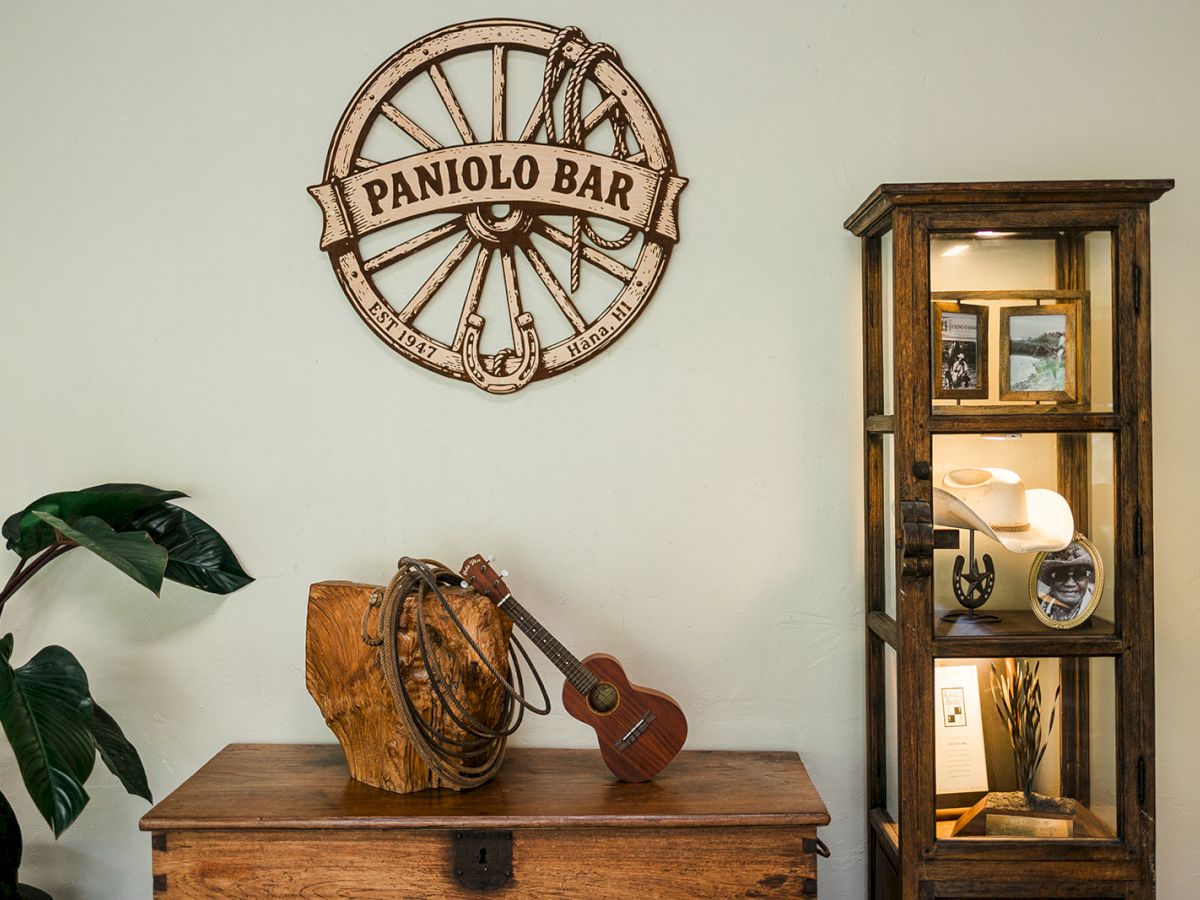 A room display with a &ldquo;Panio&rsquo;lo Bar&rdquo; wall emblem, a ukulele on a wooden table, tropical plants, and a glass-front cabinet of curios.