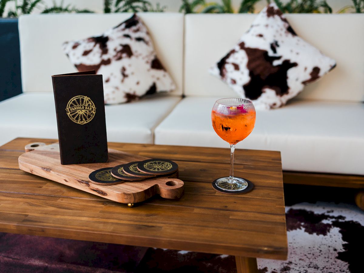 A wooden table set with a cocktail, bar menu, and coasters behind white-and-brown cow-print cushions, outdoors.