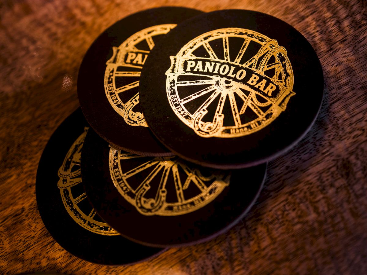 A black cap with a gold stamped emblem reading &ldquo;PANVILO BAR,&rdquo; with &ldquo;PAT&rdquo; markings, lying on a wooden table.