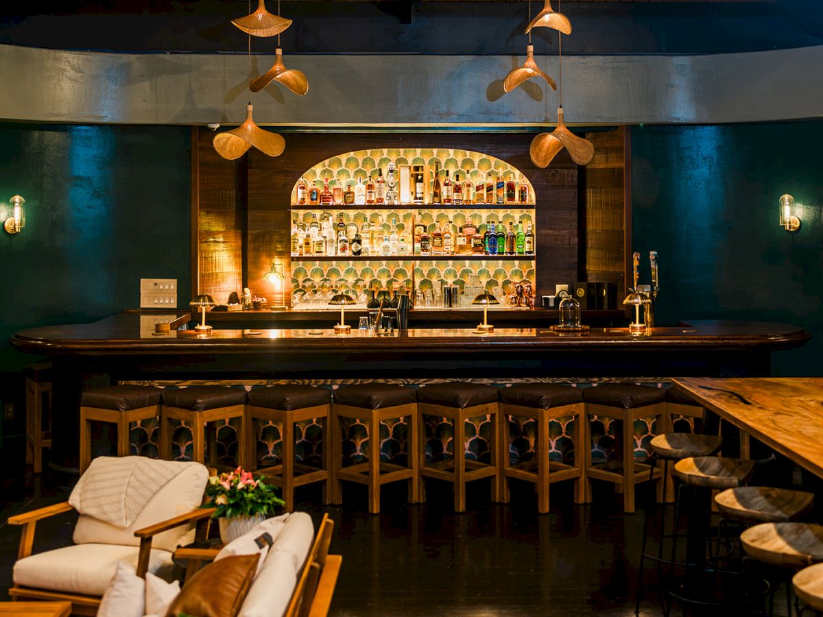 A dimly lit hotel bar with backlit liquor bottles, pendant lamps, and stools in front of a long wooden counter.