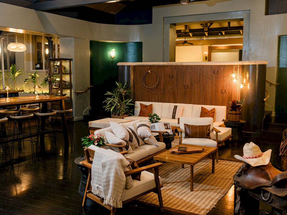A cozy indoor lounge with white sofas, wooden coffee tables, warm pendant lights, plants, and a bar-style counter area.