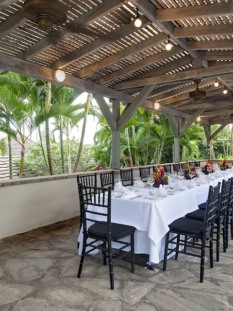 Outdoor event dining area with a long table set for guests under a wooden roof, lights and ceiling fans beside palm gardens.