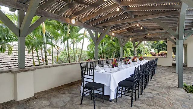 Outdoor event dining area with a long table set for guests under a wooden roof, lights and ceiling fans beside palm gardens.