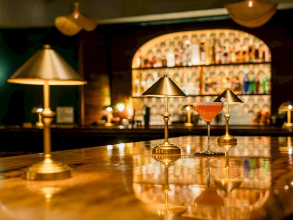 A warmly lit cocktail bar with a glowing amber drink, gold barware, and blurred bottles behind the counter.