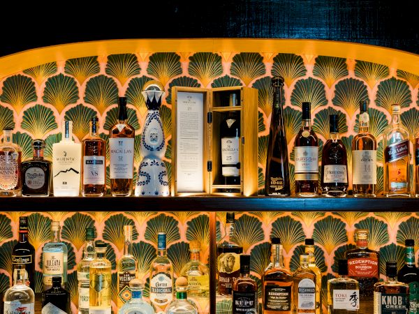 A bar shelf packed with assorted whisky, cognac, vodka, and other liquor bottles against a shell-pattern wall.