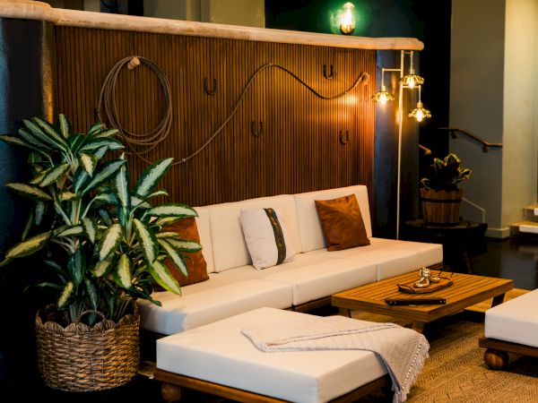 A cozy lounge area with a white sectional sofa, potted plants, a wood-slat wall, and warm hanging lights.