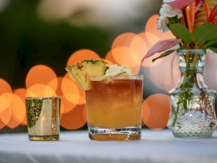 A tropical drink in a glass with pineapple garnish, set on a table with warm bokeh lights and a flower-filled vase.
