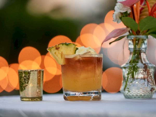 A tropical drink in a glass with pineapple garnish, set on a table with warm bokeh lights and a flower-filled vase.