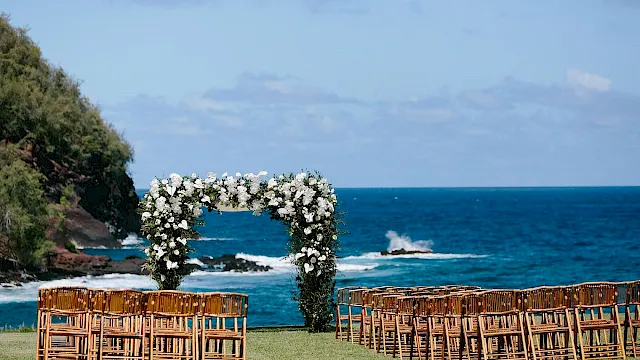 A beach wedding setup with bamboo chairs and white floral arches by crashing waves.