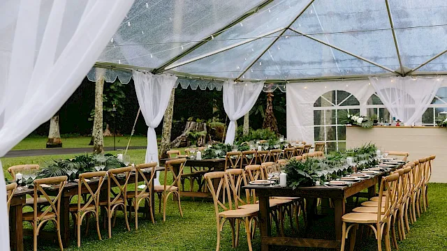 An outdoor event setup with a covered canopy, long banquet tables, wooden chairs, candles, and greenery for a catered celebration.