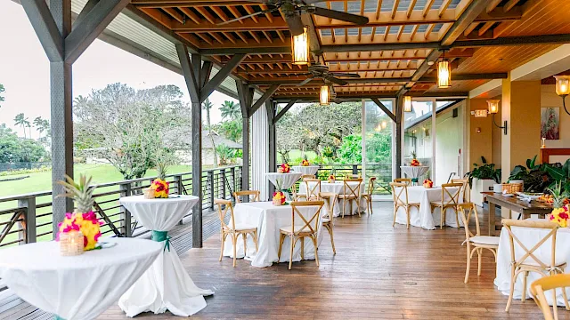 An open-air wooden pavilion set for a celebration, with dressed tables, chairs, hanging lanterns, and garden views outside.