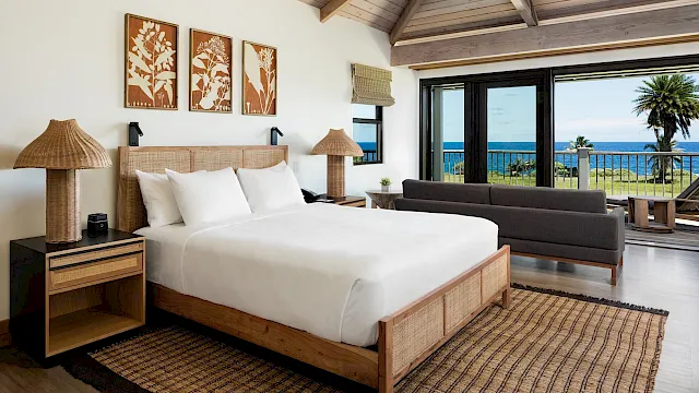 A bright seaside bedroom with a king bed, woven lamps, framed tree art, and large windows overlooking the ocean.