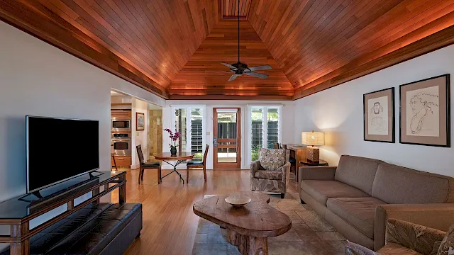 A cozy living room with a TV, sofas, coffee table, warm wood vaulted ceiling, ceiling fan, dining/kitchen area, and framed art.