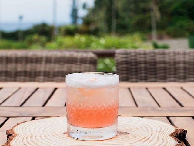 A pink-orange cocktail sits in a clear ice-filled glass on a wooden deck with palm trees and greenery in the blurry background.