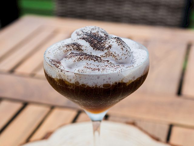 A martini glass of coffee/espresso with whipped cream and chocolate dust, on a wooden table.