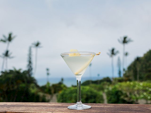 A lemon-topped cocktail in a martini glass on a wooden table with palm trees and ocean in the blurred background.