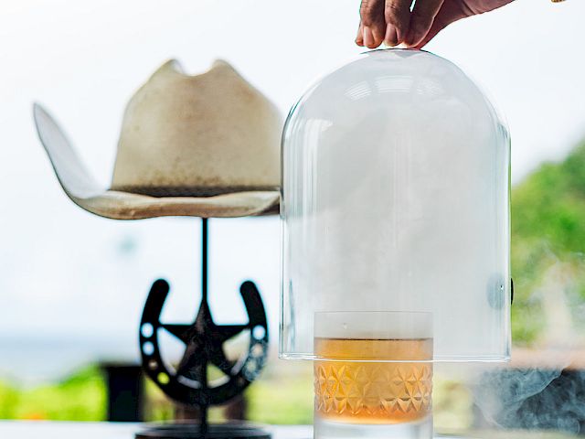 A bartender&rsquo;s hand caps a smoking cocktail with a glass cloche on a table, with outdoor decor blurred behind.