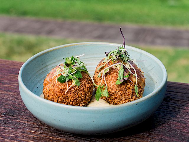 Two herb-topped meatballs served in a blue bowl on a wooden table, with a sunny garden and ocean view in soft focus behind.