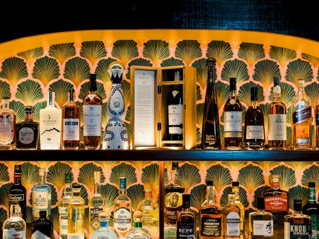 A bar shelf packed with assorted liquor bottles, including scotch and cognac, displayed against a gold seashell patterned wall.