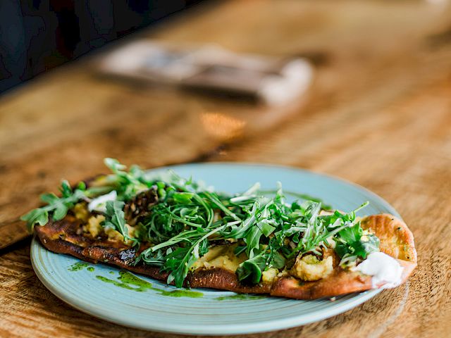 A plate of pizza/flatbread with fresh greens and cheese on a wooden table, blurred restaurant lights in the background.