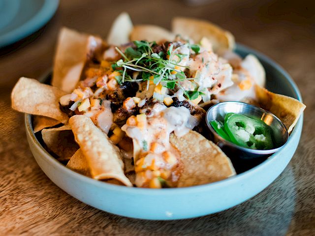 A bowl of loaded nachos with melted cheese, diced tomatoes, and cilantro, served with green salsa.