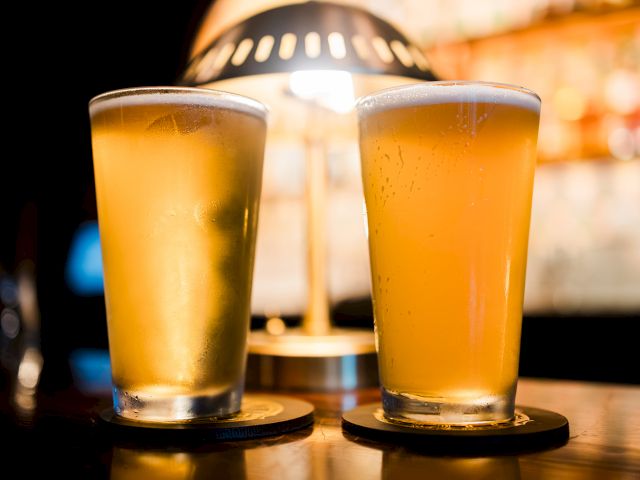 Two tall glasses of golden beer with condensation, clinking on bar coasters under warm bar lights.