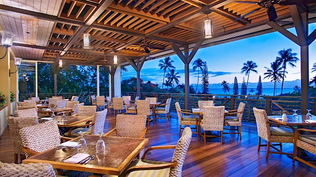 A tropical open-air dining patio with wicker chairs, lit lanterns, and a view of palm trees and the ocean at dusk.