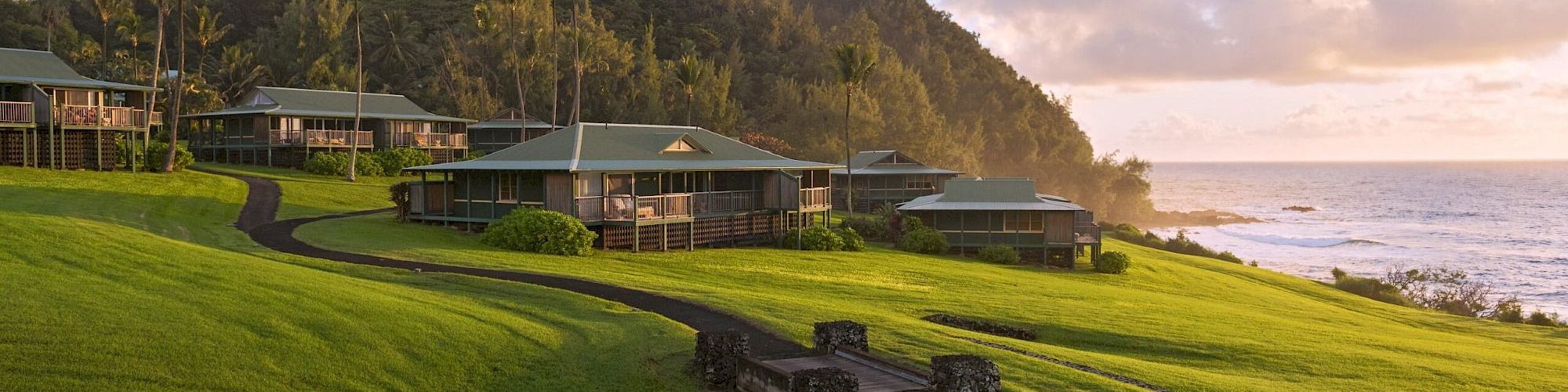 A beachfront resort with grassy slopes, palm trees, and cozy cabins overlooking the ocean beneath a cloudy sky.